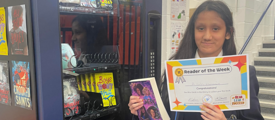 A student is in front of a book shelf holding a book and a 'reader of the week' certificate
