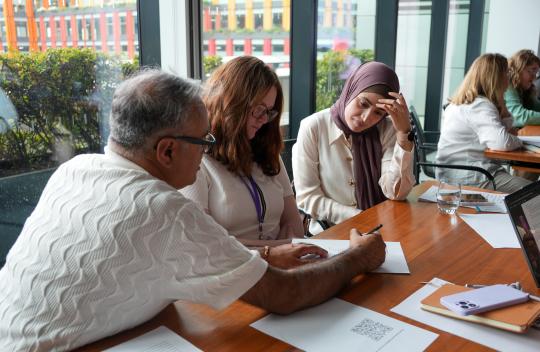 3 colleagues sit at a table looking at a piece of paper, one of them is holding a pen to write