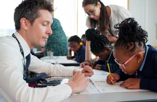 A student is sitting at a table writing in a workbook while a teacher is watching