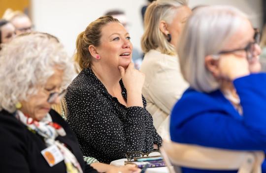 An attendee at a conference is sitting with their hand to chin listening to a speaker